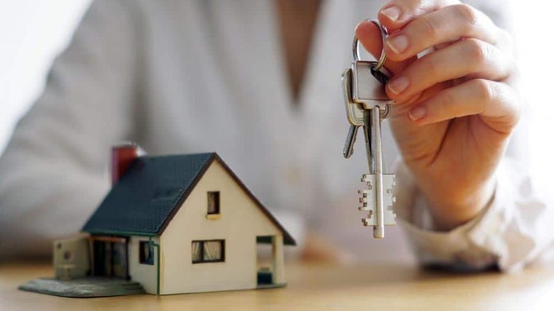 Woman Holding Keys With A Mini House Place On The Table